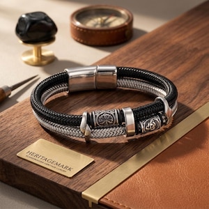 Op de afbeelding: Een dubbelstrengs armband met een zwart gevlochten koord en een zilverkleurig metalen gevlochten koord. De armband is voorzien van een gepolijste zilverkleurige magnetische sluiting en drie decoratieve metalen kralen met ingewikkelde gravures.