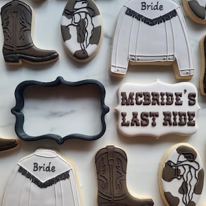 Puede incluir: Surtido de galletas de azúcar decoradas con temática western. Las galletas incluyen botas de vaquero, una chaqueta blanca con "Bride" en la espalda y galletas con el texto "McBride's Last Ride". Las galletas están decoradas con glaseado marrón, blanco y negro.
