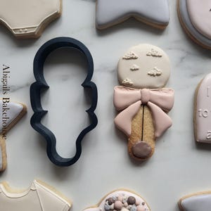 May include: An assortment of baby-themed cookies, including a onesie, star, and rattle design. The cookies are decorated with pastel colors and intricate details, perfect for a baby shower or celebration. A dark blue cookie cutter is also present.