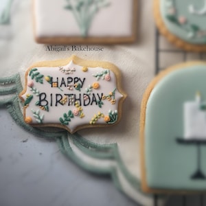 May include: A decorated sugar cookie with the words "Happy Birthday" in black icing, surrounded by floral designs in green, yellow, and pink. The cookie has a white background and a decorative edge. The text "Abigail's Bakehouse" is also visible.