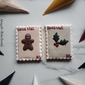 Könnte beinhalten: Zwei weihnachtliche Kekse, die wie Briefmarken gestaltet sind. Einer zeigt einen Lebkuchenmann, der andere Stechpalme. Beide tragen die Aufschrift "Santa Mail" und "30¢". Die Kekse liegen auf einer hellen Oberfläche, im Hintergrund befinden sich Spritzbeutel.