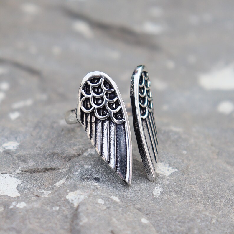 K&ouml;nnte beinhalten: Ein silberfarbener Ring mit einem offenen Fl&uuml;geldesign. Der Ring hat zwei detaillierte Fl&uuml;gel, jeder mit einem strukturierten Federmuster und einer gebogenen Form. Der Ring befindet sich vor einem grauen, strukturierten Hintergrund.