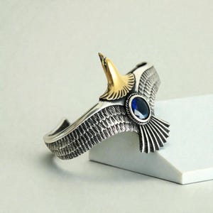 May include: Silver cuff bracelet featuring an eagle design. The eagle's head is gold-colored, and a blue gemstone is set in the center. The bracelet has detailed feather patterns and a polished finish, suitable for a bold statement.