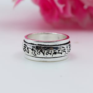 Puede incluir: Un anillo giratorio plateado con escritura negra grabada alrededor de la banda. El anillo tiene un acabado ligeramente desgastado y una banda exterior giratoria. El fondo es rosa y blanco.