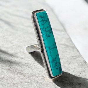 May include: A silver ring with a rectangular turquoise stone. The turquoise stone has a marbled pattern and is set in a silver bezel. The ring has a simple, modern design.