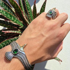 Op de afbeelding: Een zilverkleurige armband- en ringenset met een adelaarsontwerp en een groene edelsteen. De armband is een manchetstijl en de ring heeft een vergelijkbaar adelaarskopontwerp. De sieraden worden op een hand getoond.