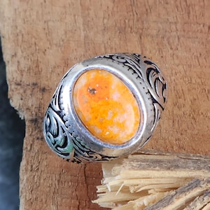 May include: Silver ring with an oval, orange-yellow stone. The ring has an ornate, engraved design around the stone and on the band. The ring is set against a rustic, wooden background.