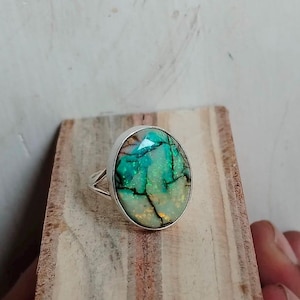 May include: Oval-shaped silver ring featuring a large, iridescent gemstone. The gemstone displays a vibrant mix of green and blue hues with dark veining. The ring is set on a wooden surface, showcasing the jewelry's design and craftsmanship.