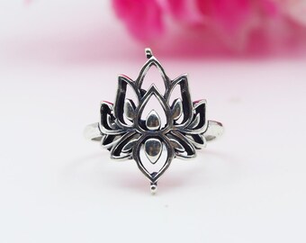 Puede incluir: Un anillo plateado con un diseño detallado de flor de loto. Los pétalos calados y los elementos centrales del anillo crean un efecto tridimensional. El anillo se encuentra sobre un fondo suave rosa y blanco, resaltando los detalles.