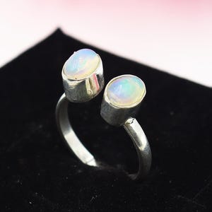 Könnte beinhalten: Ein Silberring mit zwei ovalen Opalen. Die Opale zeigen eine milchig-weiße Basis mit schillernden Farbtupfern. Der Ring hat ein schlichtes, offenes Banddesign. Die Opale sind das Hauptmerkmal des Rings.