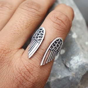 K&ouml;nnte beinhalten: Silberfarbener Ring mit offenem Design und zwei stilisierten Fl&uuml;gelformen. Die Fl&uuml;gel sind mit ge&auml;tzten Federstrukturen versehen, die eine strukturierte Optik erzeugen. Der Ring wird an einem Finger getragen.