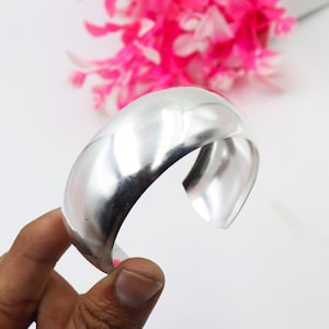 以下が含まれることがあります： 幅広で磨かれたシルバーのカフブレスレット。 オープンエンドのブレスレットは、滑らかで湾曲したデザインです。 背景には明るいピンクの花と白い表面があります。 ブレスレットは手で持たれています。