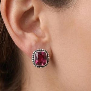 Può includere: Un orecchino a lobo con una gemma rettangolare color rosa intenso dal taglio a cuscino, incastonata in una montatura in metallo argentato. La montatura è decorata con un bordo di piccole perle argentate in rilievo attorno alla pietra.