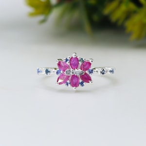 May include: A silver ring featuring a floral design. The center of the ring showcases a cluster of marquise-cut, vibrant pink gemstones, surrounded by smaller blue gemstones. Additional blue gemstones are set along the band.