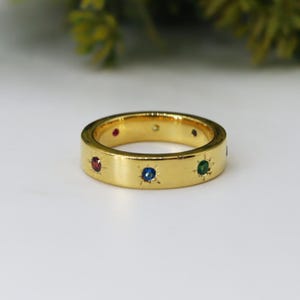 Peut inclure: Une bague dorée à bande plate, ornée de pierres précieuses colorées espacées régulièrement. Les pierres comprennent des pierres rouges, bleues et vertes, serties dans des motifs d'éclats d'étoiles. La bague est présentée sur une surface blanche.