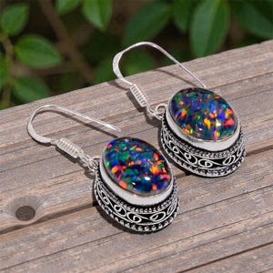 Könnte beinhalten: Silberne Ohrringe mit ovalen Opalen, die ein schimmerndes Farbspiel aus Blau, Grün und Rot zeigen. Die Opale sind in einem dekorativen, gemusterten Rahmen gefasst. Die Ohrringe haben einen Hakenverschluss.
