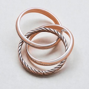 May include: Three intertwined rose gold rings. Two rings are smooth, while the third features a twisted rope design. The rings are arranged in a complex, overlapping pattern, creating a visually interesting piece of jewelry.