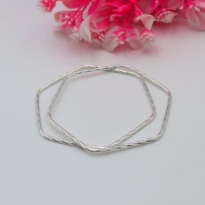 May include: Two silver-colored, geometric bangles with a textured surface are displayed. The bangles are in a hexagonal shape and are stacked on top of each other. Pink and white flowers are in the background.