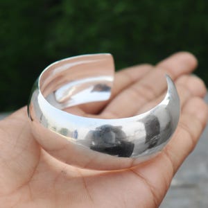 以下が含まれることがあります： 丸みを帯びたオープンデザインの、磨かれたシルバーのカフブレスレット。ブレスレットは滑らかで反射性の高い表面を持ち、開いた端は簡単に着用できます。ジュエリーは、ぼやけた緑色の背景に対して表示されています。