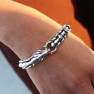 Puede incluir: Una pulsera plateada con un diseño de dragón. La pulsera está compuesta por segmentos articulados, con un cierre de cabeza de dragón. La cabeza y el cuerpo del dragón están intrincadamente detallados, mostrando escamas y una superficie texturizada. La pulsera se lleva en una muñeca.