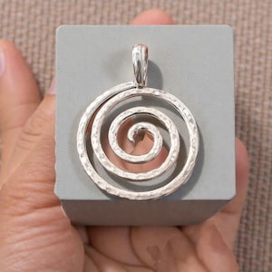Puede incluir: Colgante de plata con textura martillada, con un diseño en espiral. El colgante es circular con un lazo en la parte superior para una cadena. El diseño en espiral está centrado dentro del círculo.