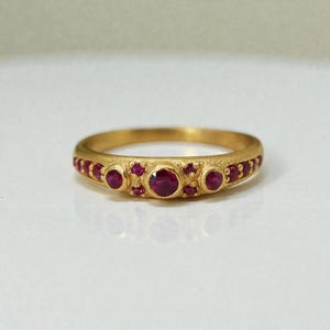 Puede incluir: Un anillo de oro con piedras preciosas de color rojo rubí. El anillo presenta un grupo central de tres rubíes más grandes, flanqueados por piedras más pequeñas. La banda tiene un aspecto texturizado y antiguo. El anillo está sobre un fondo blanco.