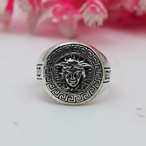 Könnte beinhalten: Silberner Ring mit einem detaillierten Medusa-Kopf-Design in der Mitte, umgeben von einem kreisförmigen Muster. Der Ring hat ein graviertes Muster am Rand und am Band. Der Stil ist klassisch und kühn. Der Hintergrund ist unscharf.