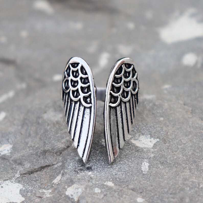 K&ouml;nnte beinhalten: Ein silberfarbener Ring mit offenem Design, der zwei stilisierte Fl&uuml;gelformen zeigt. Die Fl&uuml;gel haben detaillierte Federmuster und sind auf einer grauen Steinoberfl&auml;che angeordnet. Der Ring ist ein Schmuckst&uuml;ck.