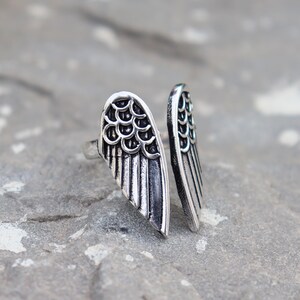 K&ouml;nnte beinhalten: Ein silberner Ring mit detailliertem Fl&uuml;geldesign. Der Ring hat zwei separate Fl&uuml;gelelemente mit Feder- und Schuppenmustern. Die Fl&uuml;gel umschlie&szlig;en den Finger. Der Ring liegt auf einer grauen Steinoberfl&auml;che.