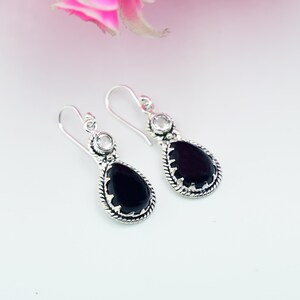 May include: A pair of silver drop earrings. Each earring has a teardrop-shaped, black gemstone, with a clear, round stone at the top. The black stone's edges are detailed with a rope-like design. The earrings are on a white background.