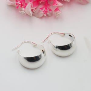 May include: A pair of silver hoop earrings with a polished finish. The earrings feature a rounded, curved design and a simple hook fastening. Pink and white flowers are in the background.