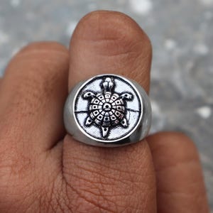 Puede incluir: Un anillo plateado con un diseño detallado de tortuga. La tortuga está centrada en una cara circular con patrones intrincados. El anillo se muestra en un dedo, destacando su tamaño y diseño.