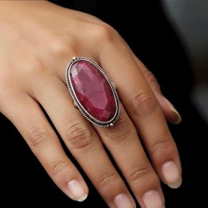 Può includere: Un anello color argento con una grande gemma ovale rosso scuro. L'anello ha un bordo dettagliato a forma di corda attorno alla pietra. L'anello è esposto su una mano.