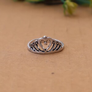 Peut inclure: Une bague en métal argenté au design inspiré d'un diadème. L'anneau présente une découpe centrale en forme de cœur surmontée d'une petite sphère, flanquée de motifs en filigrane arqués et symétriques rappelant une couronne à la finition oxydée.