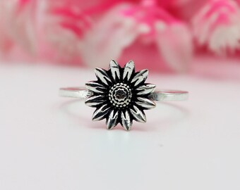 Könnte beinhalten: Silberner Ring mit detailliertem Sonnenblumen-Design. Die Blume hat mehrere Blütenblätter, die ein strukturiertes Zentrum umgeben. Der Ring ist vor einem weichen rosa und weißen Hintergrund platziert, der den floralen Schmuck hervorhebt.