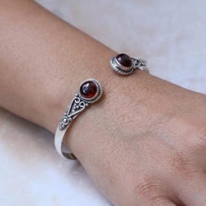 Può includere: Un bracciale a fascia in argento con due pietre granato rotonde rosso scuro fissate a ciascuna estremità aperta. La fascia in metallo presenta intricati dettagli in filigrana e piccoli accenti a perline attorno alle incastonature.