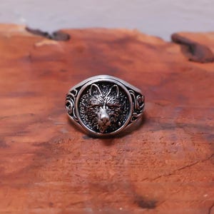 Puede incluir: Un anillo de metal plateado con una cara circular que presenta un relieve detallado de una cabeza de lobo. La banda del anillo muestra intrincados patrones de volutas que se extienden desde el motivo central del lobo hacia los lados.