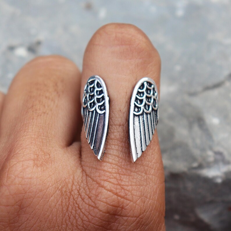 K&ouml;nnte beinhalten: Ein silberner Ring mit offenem Design, der zwei detaillierte Fl&uuml;gelformen aufweist. Die Fl&uuml;gel sind auf beiden Seiten des Fingers positioniert und schaffen ein einzigartiges Accessoire. Der Ring besteht aus gl&auml;nzendem Metall.