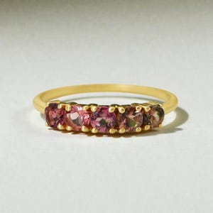 Puede incluir: Un anillo dorado con cinco gemas rosas dispuestas en fila. Las gemas presentan diferentes tonos de rosa. El anillo tiene un diseño sencillo y elegante, adecuado para el uso diario o ocasiones especiales.
