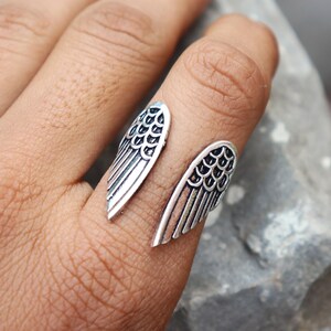 K&ouml;nnte beinhalten: Silberfarbener Ring mit offenem Design, der zwei stilisierte Fl&uuml;gelformen aufweist. Die Fl&uuml;gel haben detaillierte Federstrukturen, die ein strukturiertes Aussehen erzeugen. Der Ring wird an einem Finger getragen.