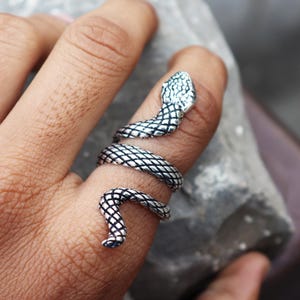 Puede incluir: Un anillo de serpiente plateado enrollado alrededor de un dedo. El anillo presenta un diseño detallado de serpiente con escamas texturizadas y una cabeza levantada. Es una joya.