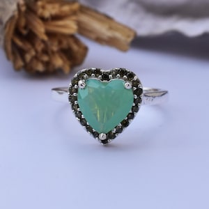 Puede incluir: Un anillo de plata con una piedra preciosa verde pálido en forma de corazón. La piedra preciosa está rodeada por un halo de pequeñas piedras oscuras. El anillo está sobre un fondo suave y neutro.