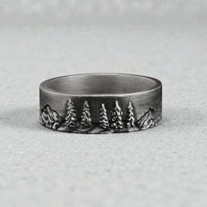 May include: A silver ring with a detailed mountain and forest design. The ring features a band with a continuous landscape of trees and mountains, creating a nature-inspired aesthetic. The ring's design is etched around the entire band.
