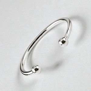 Pode incluir: Uma pulseira de punho aberta prateada, com um design liso e curvo. A pulseira apresenta uma pequena bola redonda em cada extremidade. A joia é exibida sobre um fundo branco, destacando o seu acabamento polido e a sua elegância simples.