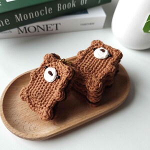 May include: Two brown crocheted bear-shaped keychains with white and black facial details sit on a wooden tray. The keychains are next to a white mug with a tea bag and a book titled "The Monocle Book of Monet."