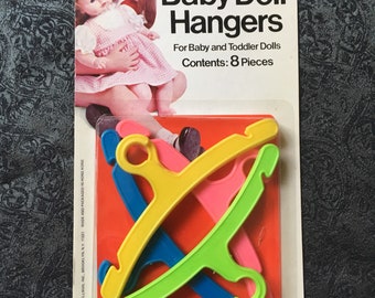 May include: A package of eight plastic baby doll hangers in yellow, blue, pink, and green. The packaging features the text Baby Doll Hangers, For Baby and Toddler Dolls, Contents: 8 Pieces, and Fits all 11 to 24 inch Dolls. The brand name Shillman is printed at the bottom.