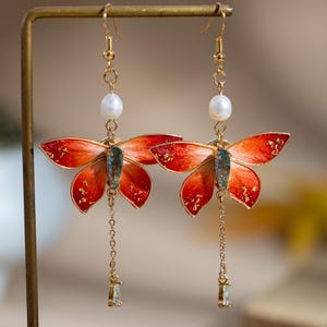 May include: A pair of gold-toned butterfly earrings. The wings are a gradient of red and orange with gold accents. Each earring features a white pearl, a blue-green gem, and a dangling chain with a small rectangular gem.