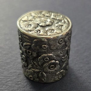 Puede incluir: Una pequeña caja cilíndrica de color plateado con un diseño floral. La tapa y los lados están cubiertos de flores en relieve. La caja es de metal y tiene un aspecto vintage o antiguo. La caja probablemente se utiliza para guardar objetos pequeños.