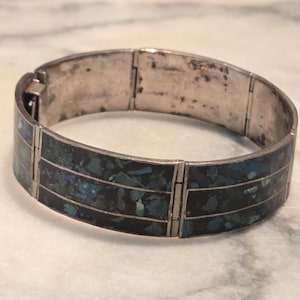 Könnte beinhalten: Ein klappbares Metallarmband mit drei horizontalen Reihen aus eingelegten, zerkleinerten Steinfragmenten in Dunkelblau, Petrol und Schwarz. Das Armband hat einen polierten, silberfarbenen Metallrahmen mit sichtbaren Scharnieren zwischen den Segmenten.