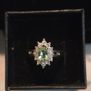 May include: A silver-toned ring featuring a central oval-cut light green gemstone surrounded by a halo of small, clear, round-cut stones. The ring is displayed inside a square black box with a velvet-like interior lining.